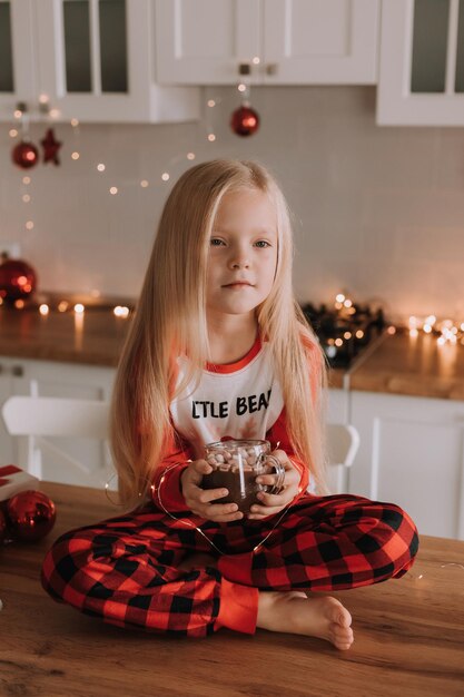 Klein blond meisje in rode kerstpyjama's met een glazen mok met een warme drank en marshmallows in een ingerichte keuken. winterse weekenden en vakanties. levensstijl. ruimte voor tekst. Hoge kwaliteit foto