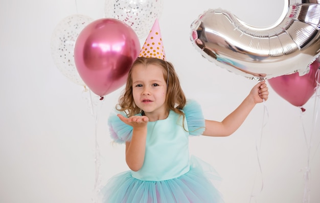 Klein blond meisje met een papieren dop houdt een bos ballonnen vast en blaast confetti uit haar hand op een witte achtergrond