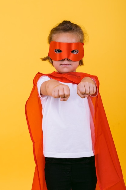 Klein blond meisje verkleed als een superheldin-superheld met een rode cape en masker, met armen in een vliegende positie, op een gele achtergrond