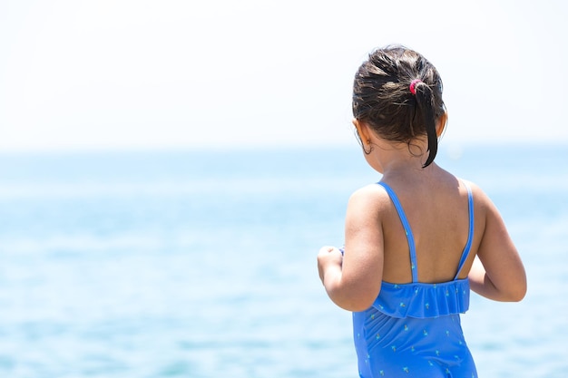 Klein brunette meisje draagt een zwembroek op het strand vanaf de achterkant met uitzicht op de oceaan met de lucht op de achtergrond Concept van strandvakantie bescherming tegen de zon zee levensstijl en ontspanning
