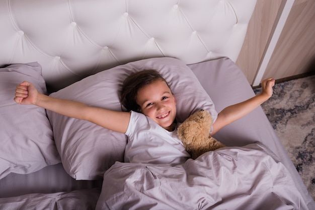 Klein brunette meisje in pyjama wordt wakker met een zacht stuk speelgoed op het bed in de slaapkamer