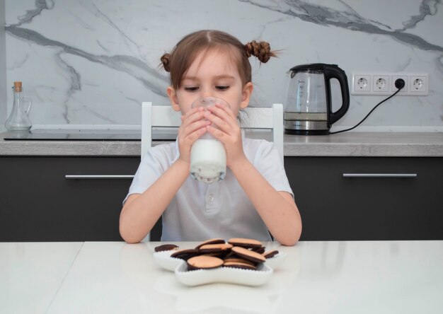 Klein charmant meisje in wit overhemd eet koekjes en drinkt melk in de keuken van het huis.