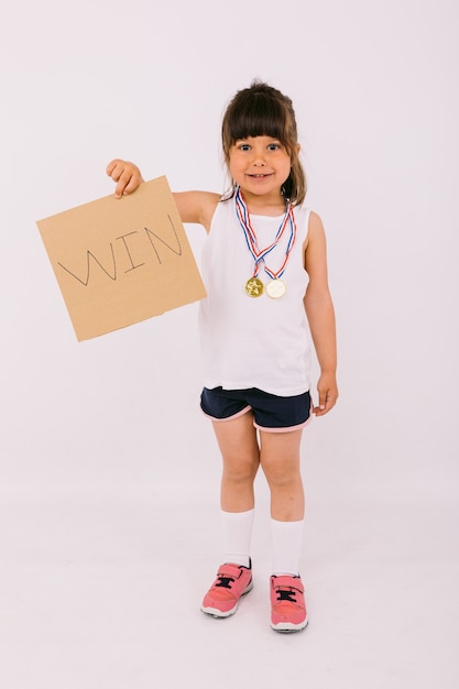 Foto klein donkerharig meisje met medailles voor sportkampioenen, met een bordje met de tekst: 'win'. sport- en overwinningsconcept