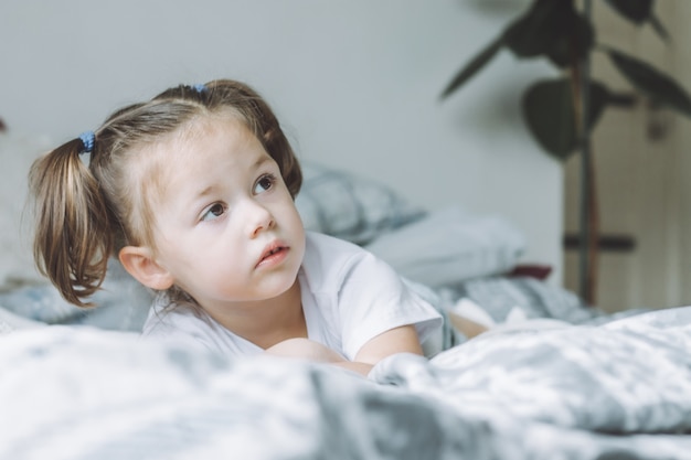 Klein donkerharig meisje met twee paardenstaarten ligt op bed op haar buik