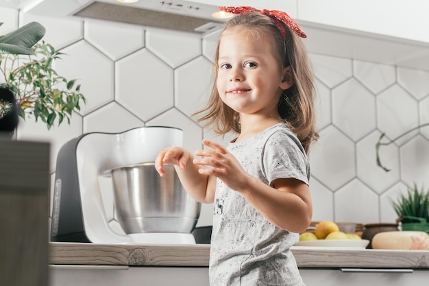 Klein donkerharig meisje van 3 jaar oud in hoofdband bakt appeltaart in de keuken. Kind staat in de buurt van planetaire mixer en glimlacht