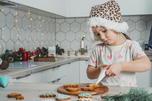 Klein donkerharig meisje van 3 jaar oud in kerstmuts versier peperkoekkoekjes met glazuur in met kerst versierde keuken