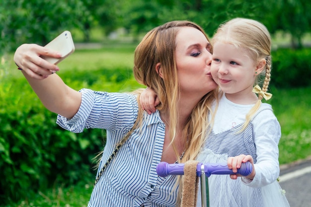 Klein en lief blond meisje in een schattige grijze jurk maakt selfie op telefoon samen met mooie moeder rust uit in het park tegen de achtergrond van bomen en groen