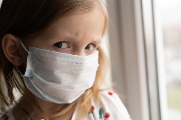 Klein Europees meisje draagt masker ter bescherming van Covid-19. Ziek kind Kleine meisjes kijken naar het raam. Kopieer ruimte