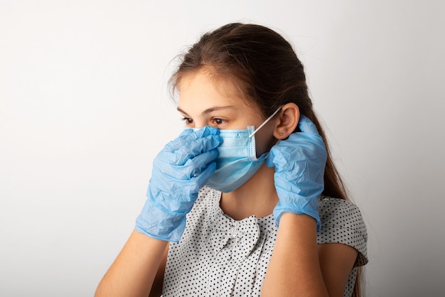 Klein Kaukasisch meisje in medisch masker en handschoenen in studio. Schoolmeisje in jurk zet op een beschermend medisch masker