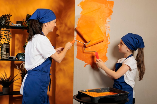 Klein kind helpt moeder om muren te schilderen, oranje verf te gebruiken om de huiskamer te renoveren en opnieuw in te richten. Jong gezin renoveren appartement met penseelroller en gereedschap, huishoudelijk werk.
