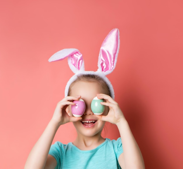 Klein kind in hoofdband met konijnenoren blauwe tshirt ze lachte haar ogen bedekt met eieren poseren op roze achtergrond pasen