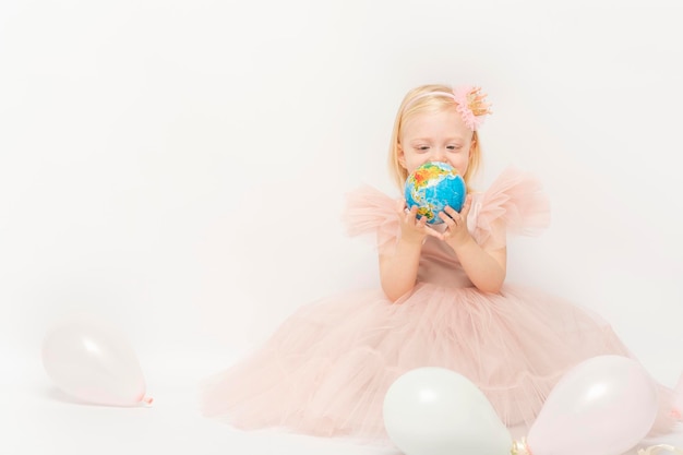 Klein kind kijkt naar kleine wereldbol in handen Blond meisje in jurk met tule rok zit op witte achtergrond met ballonnen