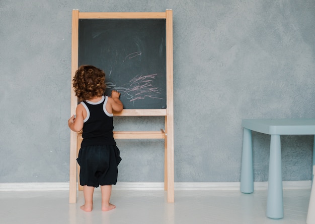 Klein kind tekent thuis met krijt op een zwart schoolbord in de kinderkamer tegen een grijze muur.