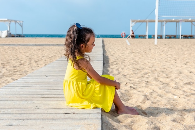 Klein krullend meisje in een gele jurk zit op een verlaten zandstrand op een zonnige dag, leuke vakanties