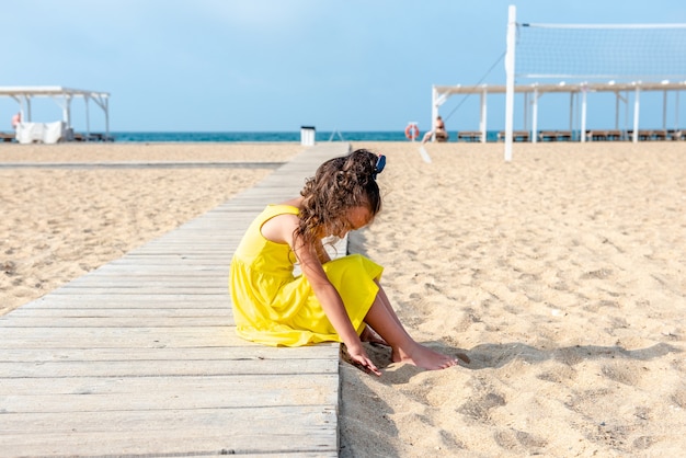 Klein krullend meisje in een gele jurk zit op een verlaten zandstrand op een zonnige dag, leuke vakanties