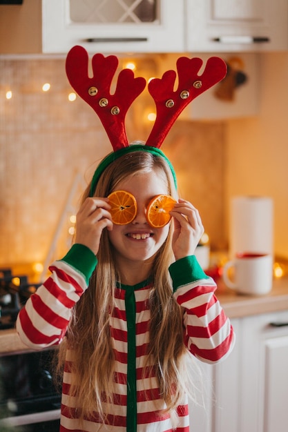 Klein lachend schattig blond meisje in pyjama en gewei houdt fruithelften in de buurt van haar ogen. kind in de keuken bij de kerstboom. Kerstmis