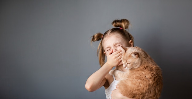 Klein meisje 7-8 jaar oud met een rode kat in haar armen lijdt aan allergieën, niest door een allergische rhinitis, grijze muur