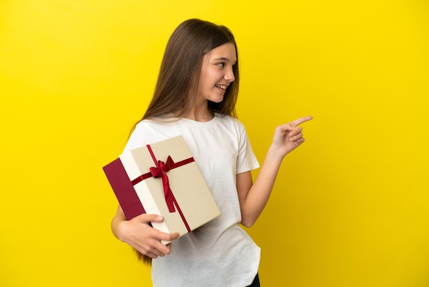 Klein meisje dat een cadeau vasthoudt over een geïsoleerde gele achtergrond die met de vinger naar de zijkant wijst en een product presenteert