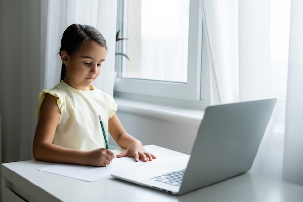 Klein meisje dat naar het laptopscherm kijkt met een uitdrukking van verrast en opgewonden. Slim, glimlachend meisje dat aantekeningen maakt. Communicatie in bedrijfsconcept.