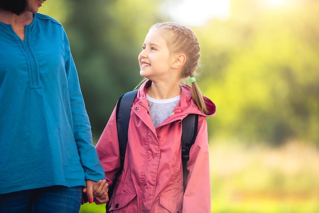 Klein meisje dat naar school gaat met moeder die hand vasthoudt en lacht op zonnige natuurachtergrond