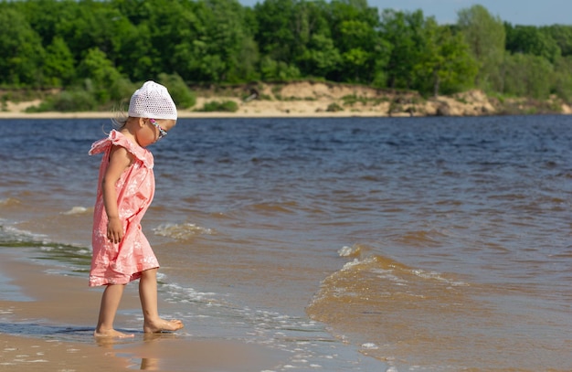 Klein meisje dat op het zomerstrand loopt, gewassen door golven met een prachtig landschap op de achtergrond