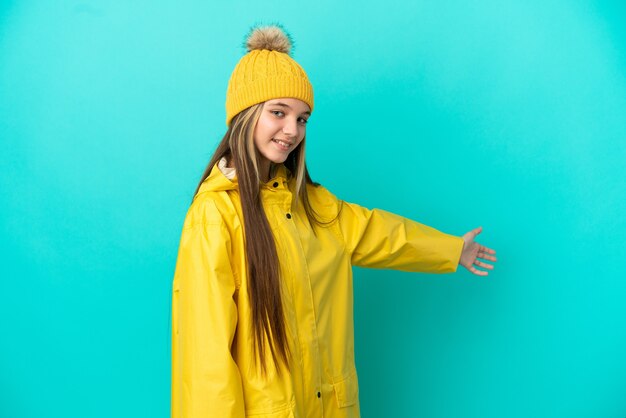 Klein meisje draagt een regenbestendige jas over een geïsoleerde blauwe achtergrond die haar handen naar de zijkant uitstrekt om uit te nodigen om te komen