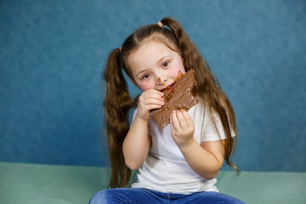 Klein meisje eet chocolade en veegt haar witte t-shirt, gezicht en geeft hem