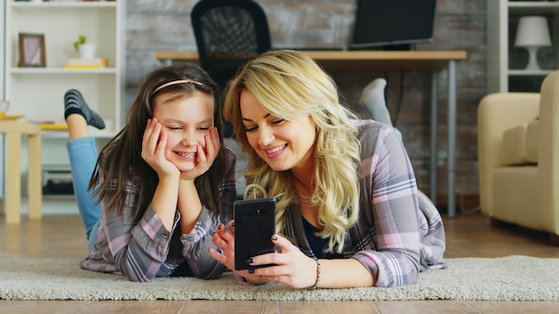 Klein meisje en haar moeder liggend op de vloer met smartphone om te winkelen.