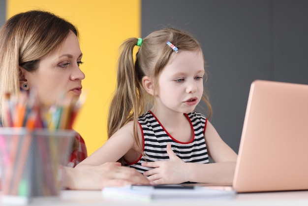 Klein meisje en moeder zitten aan tafel voor het laptopscherm