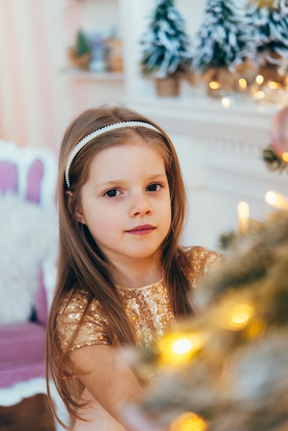 klein meisje, gekleed in een mooie modieuze witte jurk, poseert in de buurt van de kerstboom.