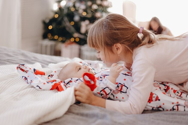 Klein meisje gekleed in pyjama kijkt naar haar kleine broertje liggend op het bed in de gezellige kamer met nieuwjaarsboom.