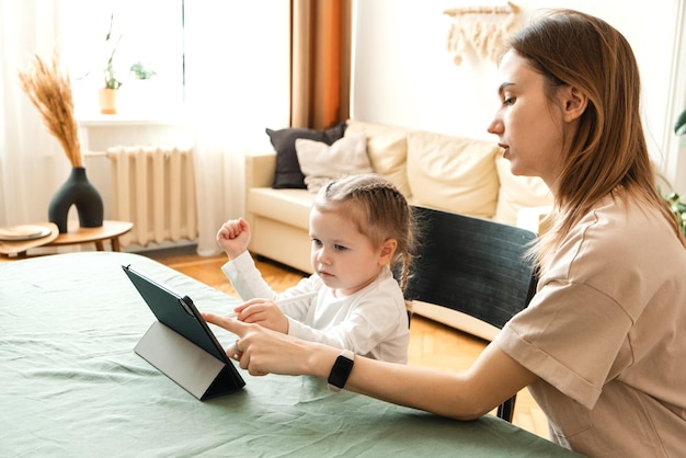 Klein meisje heeft moeders hulp nodig bij het doen van taken op de tablet thuis Moeder helpt haar dochter haar huiswerk te maken voor de kleuterschool