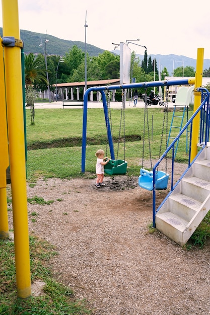 Klein meisje houdt de schommel vast met haar handen op de achtergrond van gebouwen en park