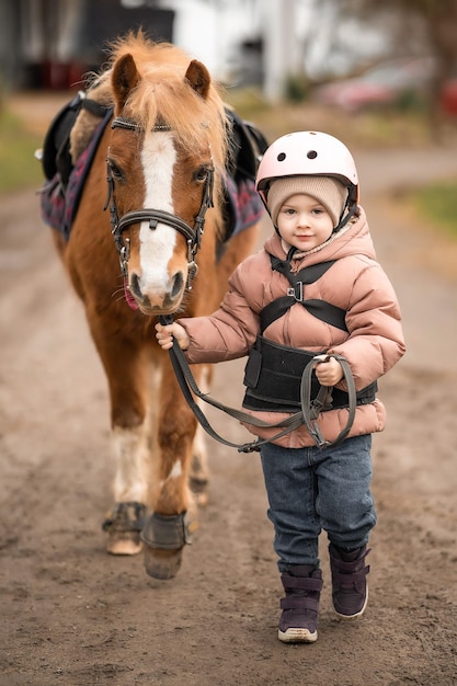 Klein meisje in beschermende jas en helm met haar bruine pony voor rijles