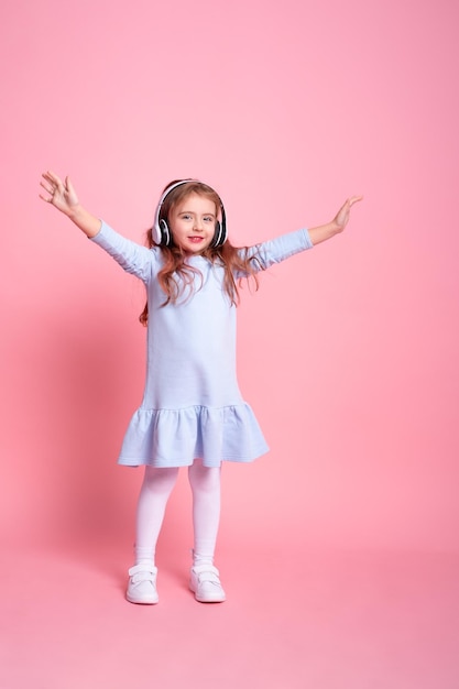 Klein meisje in blauwe jurk luisteren naar muziek dansen in grote witte koptelefoon op roze achtergrond