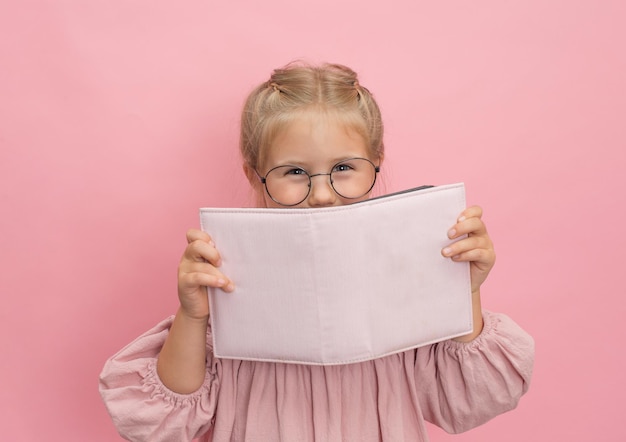 Klein meisje in bril met boekalbum of dagboek op roze achtergrond