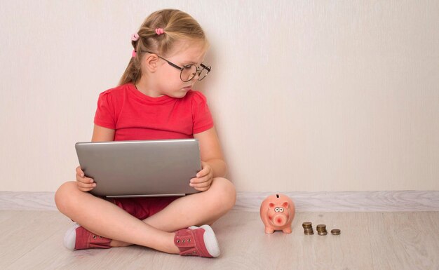 Klein meisje in brillen met behulp van notebook laptop zittend op de vloer met spaarpot en munten