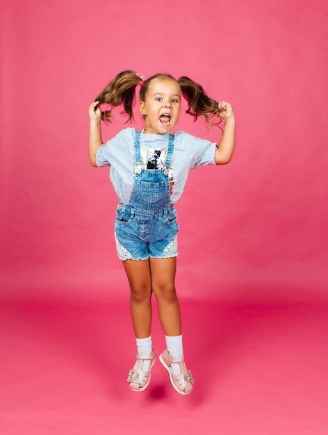 Klein meisje in denim overall springen op een roze achtergrond. vrolijk kind. kinderen.