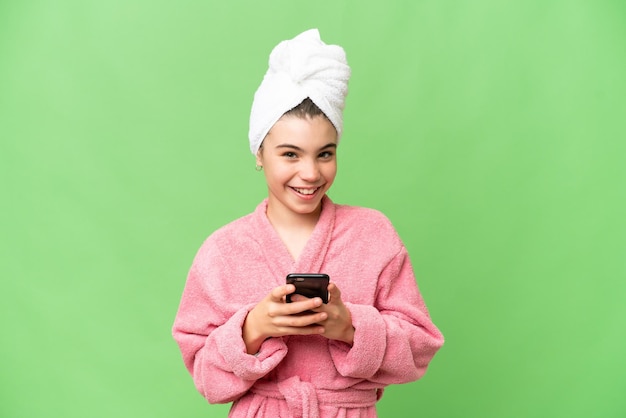 Klein meisje in een badjas over geïsoleerde chroma key-achtergrond die een bericht verzendt met de mobiel