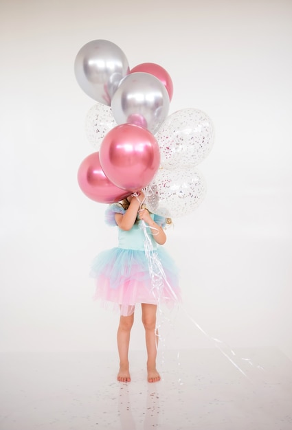 Klein meisje in een feestelijke jurk met een tutu-rok staat met een bos ballonnen en bedekte haar gezicht op een witte achtergrond