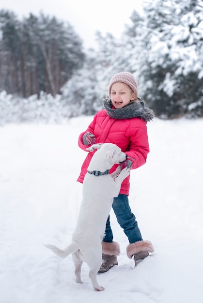 Klein meisje in een fel jasje speelt in het besneeuwde winterbos met haar hond jack russell terrier