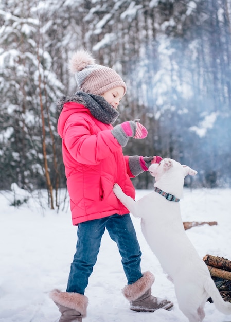Klein meisje in een fel jasje speelt in het besneeuwde winterbos met haar hond jack russell terrier