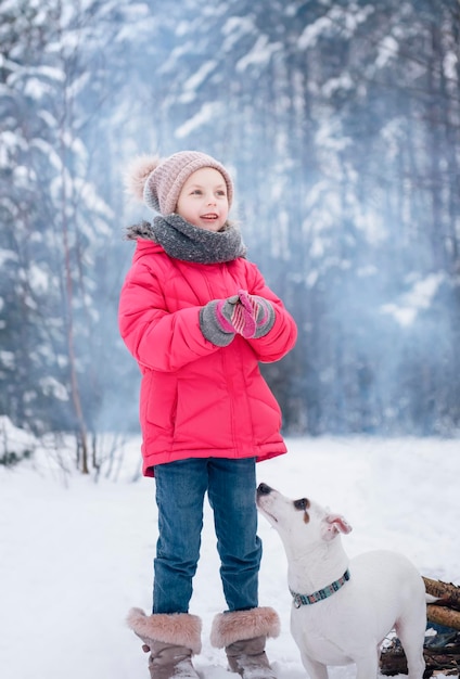 Klein meisje in een fel jasje speelt in het besneeuwde winterbos met haar hond jack russell terrier