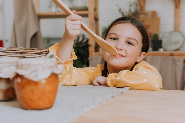 klein meisje in een gele jurk met een houten spatel in haar handen en jam in glazen pot in de keuken