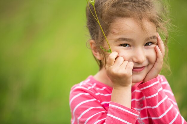 Klein meisje in een koraal gestreept T-shirt op een groene achtergrond in een veld houdt haar gezicht in haar handen en glimlacht sluw. Kinderdag, blij kind, milieu- en natuurbescherming, insectenwerend middel