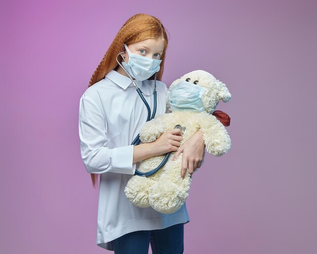 klein meisje in een medisch masker luistert naar een teddybeer door middel van een stethoscoop covid preventie concept