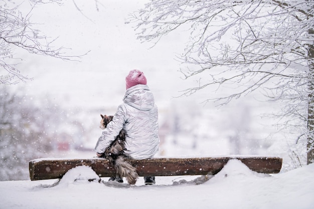 Klein meisje in een omhelzing met een grote kat die met haar rug naar de kijker op een bank zit te kijken naar het met sneeuw bedekte landschap dat zich vanaf de heuvel ontvouwt.