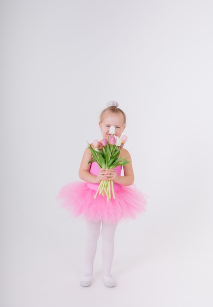 Klein meisje in een roze jurk met een tutu rok staat met een boeket tulpen op een witte muur