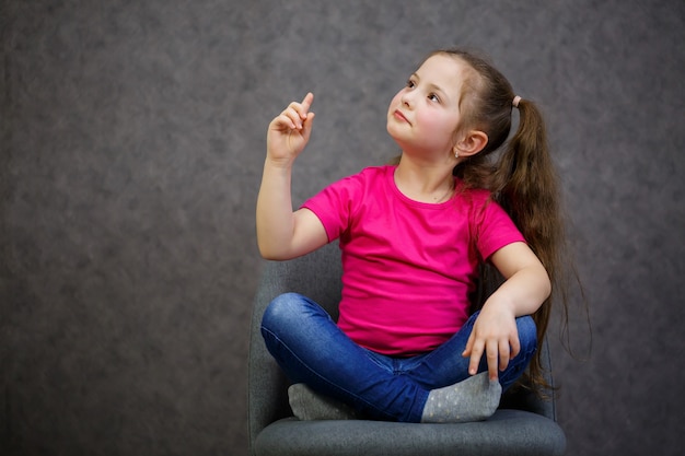 Klein meisje in een roze t-shirt zit op een grijze stoel. Emotionele foto van een kind