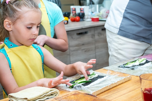 klein meisje in een schort bereidt sushi voor door komkommers te leggen